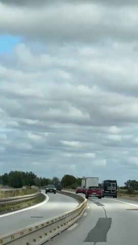 The video shows a view from a vehicle on a highway. Several cars and trucks are driving on the road. The sky is cloudy. In the distance, on the left side of the highway, a police car with flashing lights is visible. It appears to be stopped or moving slowly in a way that indicates it is interacting with another vehicle or situation.