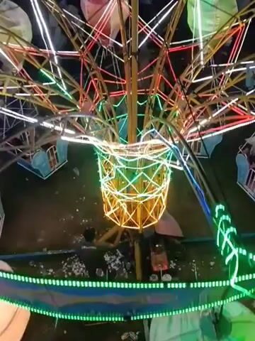 The video shows an overhead view of a carnival ride with multiple gondolas suspended from a central structure. The ride is illuminated with colorful lights, primarily green and yellow. Several gondolas are visible, each appearing to be designed to resemble a hot air balloon basket. People are seated in some of the gondolas. One gondola in the foreground, directly below the main illuminated structure, shows two individuals. Another gondola to the right has several people inside. The ground below the ride is dark, and some debris is scattered on the ground. The ride appears to be in motion, as the gondolas are swaying.