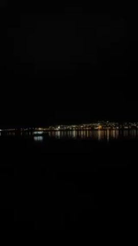 At night, a body of water reflects the lights of a distant city. A small boat sits still in the water in the foreground. The sky is dark and featureless.