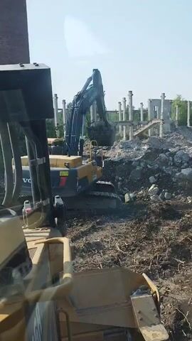A demolition site with two excavators and a bulldozer. One excavator is yellow and black with "VOLVO" on the side. It is in the foreground to the right. The second excavator is black and in the background, lifting debris. A bulldozer is in the immediate foreground, partially obscured by the camera's perspective. Rubble and dirt are scattered around the site. The sky is visible overhead.