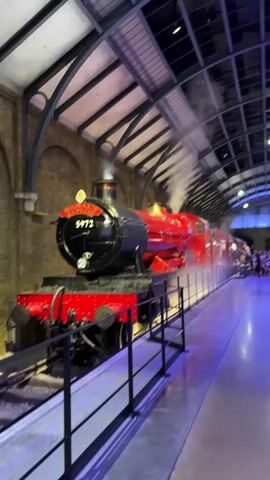 This video shows a platform at a train station, likely a recreation of King's Cross Station from the Harry Potter series. A red steam engine, labeled "9 1/2", is prominent on the left side, emitting steam. The platform is lined with people, many of whom appear to be tourists, some taking photos or waiting. The station has a vaulted ceiling with industrial lighting and brick walls. The floor is polished concrete, reflecting the blue and purple lighting that casts a dramatic ambiance over the scene. Several signs on the wall, including one that says "HOGWARTS EXPRESS", confirm the Harry Potter theme. People are seen walking along the platform, interacting with the environment, and some are pushing luggage. The overall atmosphere is one of excitement and wonder, particularly for fans of the Harry Potter franchise.