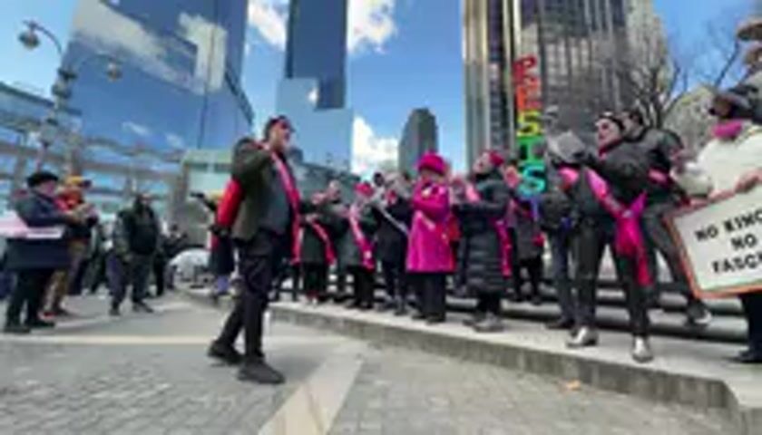 A street-level video captures a demonstration or protest in an urban setting. In the foreground, a man wearing a dark jacket, black pants, and a red backpack, with a pink sash draped across his shoulder, is actively engaging with a group of people. He holds a microphone close to his mouth and appears to be leading a song or chant, stepping and gesturing enthusiastically.

Behind him, a group of approximately fifteen to twenty individuals, mostly women, are also participating. Many of them are wearing pink sashes, some with pink hats or coats, and are clapping or making hand gestures in rhythm with the singing. One person on the far right holds a white sign that clearly reads "NO KINGS NO FASCISM" in black block letters. Another sign, partially visible in the background, shows letters that appear to spell "RESISTS" in green.

The background features tall, modern glass-fronted buildings under a clear blue sky with some scattered clouds, indicating a bright, sunny day. The ground is paved with light-colored cobblestones or patterned concrete. The participants are singing in unison, and snippets of their song can be heard: "...just how badly we failed. Don't you wish we were dreaming. Is this the level end... will never end." The overall atmosphere suggests an organized gathering with a clear political or social message.