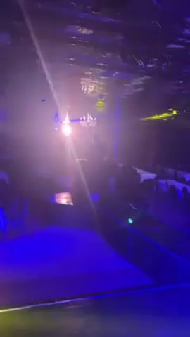 The video shows a dimly lit room, possibly a ballroom or event space, with blue and white lights adding to the ambience. Rows of tables and chairs are visible, suggesting it is set up for a formal event. In the foreground, a bright light shines directly into the camera, creating a lens flare effect. As the camera pans, three women come into view dancing energetically, their forms blurred by motion. One woman wears a black top and a vibrant red skirt, another is in a black top and blue jeans, while the third has on a light colored dress. The focus is on their dancing, with the background details remaining somewhat indistinct.