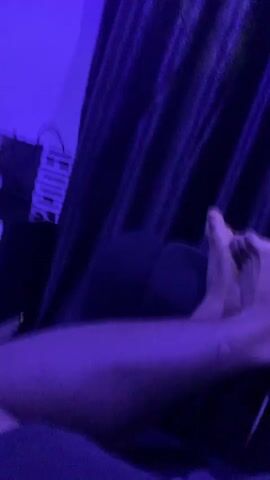 The video begins with a close-up of a person's bare feet and lower legs. The scene is dimly lit and bathed in purple and blue light, possibly from a LED light source. The person's feet appear to be resting on a dark surface, possibly a bed or sofa, with a black curtain visible in the background. As the video progresses, the camera pans to the right, revealing a shelf unit with various objects on it. These objects include books, what appears to be a gaming console, and other miscellaneous items. The lighting remains consistent throughout the clip, casting shadows and creating a moody atmosphere. A sign with the letters "CODE" is visible on the shelf unit. The camera movement is somewhat shaky, suggesting it is being held by hand. The overall impression is of a casual, dimly lit indoor setting, possibly a bedroom or living room, with a focus on the feet and then a wider view of the surroundings.