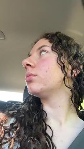 A person with curly brown hair is riding in the passenger seat of a car and looking out the window. A man is driving the car and wearing sunglasses. The car is on a road surrounded by green trees.