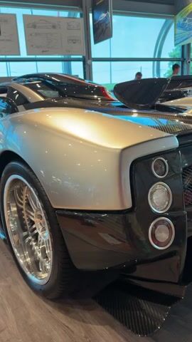 The video shows a close-up of the rear of a silver and black luxury sports car, likely a Pagani. The car is parked indoors, possibly in a showroom, with large windows and display posters visible in the background. The camera focuses on the car's taillights, which are circular and stacked vertically, and the large rear spoiler. The car's bodywork features a metallic silver finish on the fender and a glossy black finish on the lower sections and rear diffuser. A part of the car's wheel with chrome spokes is visible on the left. The overall impression is one of high-end automotive design and engineering.