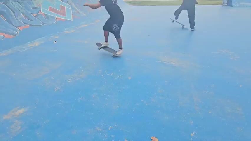 The video shows two young men skateboarding in an outdoor skate park. One skater is in the foreground wearing a black t-shirt and dark shorts, bending low with his arms out for balance as he rides across a blue concrete surface. Another skater in dark clothing is in the background, further away from the camera, also skateboarding on the same blue surface. Graffiti art is visible on the side of a ramp in the background to the left, and a metal rail is also in the background to the right. The surface of the concrete is worn and shows signs of use. The overall lighting indicates a bright, sunny day.
