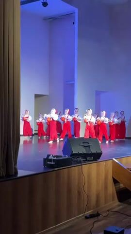 A short clip shows a group of about ten young girls in red and white costumes performing a dance on a stage. They are holding what appear to be dolls or toys. The stage has a dark floor and is lit with blue light. A large speaker is positioned on the front edge of the stage. To the left, a curtain is partially drawn. The background consists of white walls with tall, rectangular architectural features.