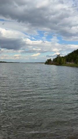 The video shows a calm lake with a slightly rippled surface, reflecting a cloudy sky with patches of blue. The camera pans slowly to the right, revealing a wooded shoreline. The trees are dense and green, with some rocky outcrops visible. The water is clear enough to see the sandy bottom near the shore. The overall impression is one of a peaceful, natural landscape.