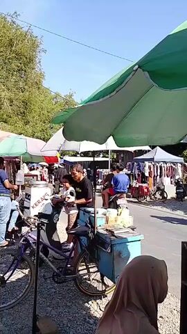 The video shows a bustling outdoor market scene on a sunny day. In the foreground, a person wearing a light-colored cap and a dark shirt is shown preparing food in a large wok under a green umbrella. The wok is filled with small, white food items being fried. To the left, a purple bicycle is parked. In the middle ground, several market stalls are visible, with people browsing and interacting. One stall displays clothing on racks. A few motorcycles are parked nearby. Further in the background, more market stalls, trees, and buildings can be seen, along with more parked motorcycles and people walking around. The overall atmosphere appears lively and active, typical of a street market.
