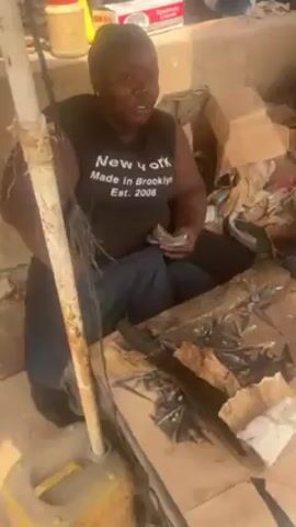 The video depicts a bustling outdoor market setting in an unspecified location. Two women are prominent in the initial frames, sitting amidst scattered debris, including cardboard boxes, plastic bags, and what appear to be dried fish. One woman, wearing a black tank top that reads "New York Made in Brooklyn Est. 2008," is counting a wad of cash. The other woman, donning an orange shirt and a pink head covering, appears to be sorting through items near a cell phone. As the camera pans, the scene reveals a crowded market, with various vendors and customers moving about. The market structure is rudimentary, with a makeshift roof providing shade. The overall impression is of a vibrant, active commercial environment in a developing area.