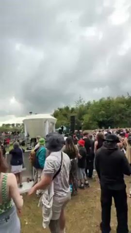 Here's a description of the video:

The video shows a crowd of people at what appears to be an outdoor music festival. The camera pans across the scene, capturing the audience and the stage in the background. A DJ setup is visible on the stage. The people in the crowd are wearing various types of clothing, and some are dancing. A flag is visible in the background, and the sky is cloudy. Overall, the video captures a lively and energetic atmosphere at the festival.