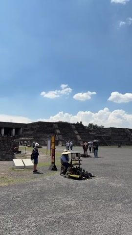 The video shows the Pyramid of the Sun at Teotihuacan, Mexico.  The sky is blue with puffy white clouds. The pyramid dominates the view, with people climbing its steps and walking around its base. Several vendors are present, displaying their goods on small carts. The ground is gravel. People are casually dressed, and the overall atmosphere appears to be relaxed and touristy.