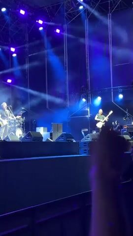 A live band is performing on a stage with a large audience in front of them. The stage is lit with blue and purple lights and there is smoke in the air. Two guitarists are visible on stage, one in the left foreground playing a guitar, and another in the right foreground also playing a guitar. A drummer is visible behind the guitarist on the left. A hand is in the foreground of the shot, obscuring part of the view of the stage. The audience is not clearly visible, but there are some people at the bottom of the frame, suggesting a large crowd.