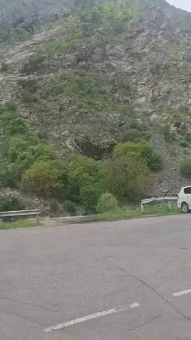 The camera pans up from a view of a paved road and guard rail to show a hillside with a mix of exposed rock and green trees. The hillside rises steeply. Power lines are visible running across the sky above it. A small white building is momentarily visible in the lower left. The sky is overcast.