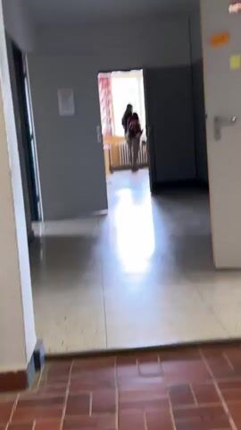 The video begins with a shot of a bright, reflective surface, possibly a floor or a mirror. The perspective then shifts to show a person standing in a classroom. They are wearing a black t-shirt, grey pants, and dark shoes, and are holding a backpack with colorful accents. The classroom has a radiator along the wall and a window with curtains. The video then pans to show a hallway with open doors. A person, similar in appearance to the first, is visible through an open doorway, stooping over. The environment appears to be a school or similar institution.