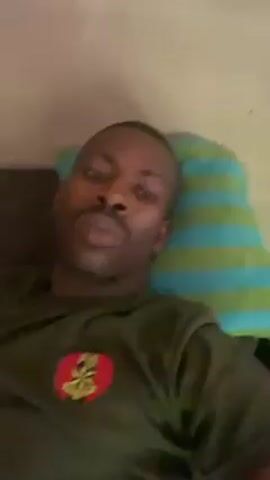 A man with dark skin, close-cut hair, and a mustache is lying on his back. He appears to be on a couch or bed with a striped pillow behind his head. He is wearing an olive green t-shirt with a circular emblem on the chest. His eyes are closed initially, then he opens them and looks directly at the camera. The background is a plain, off-white wall. The camera appears to be held by the man, giving a first-person perspective.