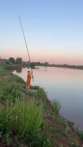 A young boy wearing an orange shirt and shorts is standing on the grassy bank of a calm lake, holding a long fishing rod. He has just cast the line into the water, creating ripples. He is standing on a purple mat on the dirt and grass edge of the bank. The sky is a pale blue and pink, indicating dawn or dusk. In the background, there are trees, a small outbuilding, and a red-roofed structure, suggesting a rural setting. The grass on the bank is lush and green.