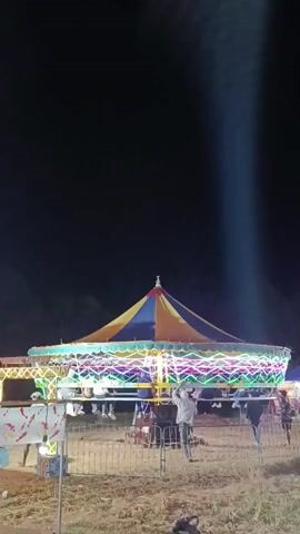 The video opens with a shot of a brightly lit carousel at a night fair. The carousel has a colorful canopy and is decorated with various lights. Several people are visible on and around the carousel, some appearing to be riding it while others watch. The camera then pans to the right, revealing a Ferris wheel in the background, also illuminated with lights. A group of people, mostly women wearing headscarves, are standing in the foreground, facing away from the camera and looking towards the rides. The ground appears to be uneven and grassy. The overall atmosphere suggests a lively and festive event.
