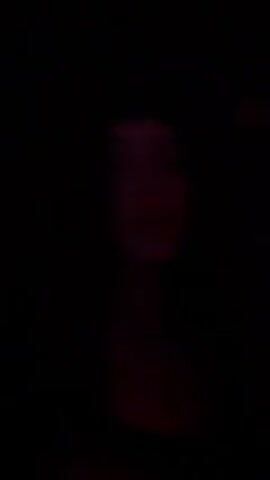 A young woman with long curly hair, wearing a black headband and a necklace with a cross pendant, is speaking in a dimly lit environment with red lighting. She looks directly at the camera. The background is dark and out of focus. The sound of music is audible.