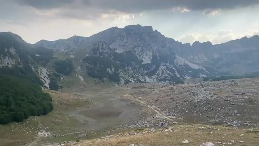 The video shows a panoramic view of a mountainous landscape under a cloudy sky. The sun is shining through the clouds, creating rays of light that illuminate the scene. The foreground is covered with dry grass and scattered rocks. In the middle ground, there is a wide valley with a dry lakebed or pond in the center, and a dirt road winding through it. The background features a range of rugged mountains, some with steep, rocky slopes and others covered with patches of green vegetation. To the left, there is a dense forest of green trees. The overall impression is one of a vast, natural, and somewhat desolate wilderness. There are no people or animals visible in the video. The camera pans slowly across the landscape, offering a sweeping view of the terrain and the interplay of light and shadow.