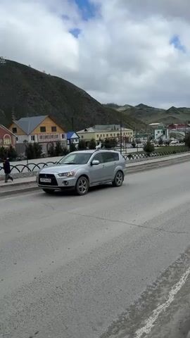 The video shows a street in a mountainous area, with various cars passing by. In the background, there are several buildings, and a mountain with what appears to be an inscription on it. The sky is partly cloudy. The road itself is gray and has some visible wear. A silver SUV is parked on the left side of the road at the start, followed by a dirty white SUV passing by. The road curves slightly as it continues into the distance. A fence, with a recurring semi-circular pattern, separates the road from the buildings.