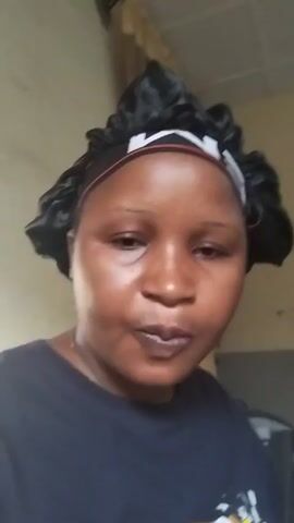 A woman is holding up a plate of cubed food to the camera. She wears a black hair bonnet with a white pattern and a black t-shirt. She is smiling and talking. The background shows a wall painted off-white and white-tiled floors.
