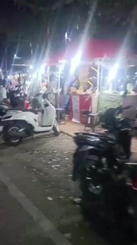 The video opens showing a white scooter parked on the side of a road at night. People are visible in the background, with stalls set up and lit by string lights. More scooters are parked in front of the stalls, and people are gathered around them. The camera pans slightly to the left, revealing more of the busy scene. The lighting is dim, with bright lights from the stalls illuminating the area. The ground appears to be a mix of dirt and pavement. The overall impression is of a lively night market or roadside gathering.