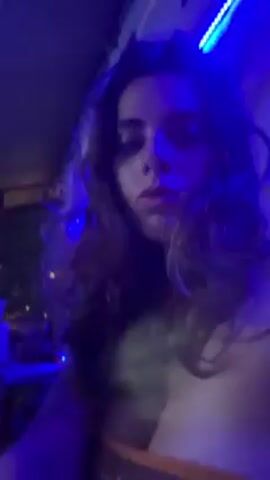 The video shows a young woman standing in a dimly lit bar or club. She has long brown hair and is wearing a top. Behind her is a pool table with several balls on it. People can be seen playing pool in the background. The lighting in the room is changing colors, shifting between blue, purple, and red. The woman is looking at the camera and appears to be recording a video of herself.
