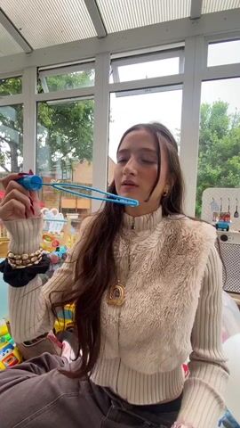 A young woman with long brown hair, wearing a beige fur vest over a beige ribbed sweater and dark pants, is seated indoors, blowing bubbles with a blue bubble wand. She holds the wand in her right hand, bringing it to her mouth, then lowers it and smiles at the camera. Her left wrist has several bracelets and a black scrunchie. The background shows a room with large windows, revealing trees and a small playhouse outside. Inside, there are various children's toys and balloons scattered around.