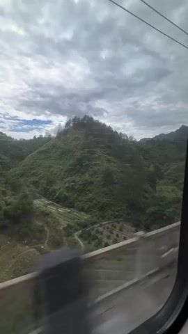 The video shows a view from a moving train window, looking out at a landscape of green hills and cloudy skies. The train is traveling alongside a concrete barrier, with lush vegetation between the barrier and the hills. The sky is overcast with a mix of gray and white clouds. In the distance, the rolling hills are covered in dense trees and vegetation. The overall scene is one of a journey through a rural, natural environment.