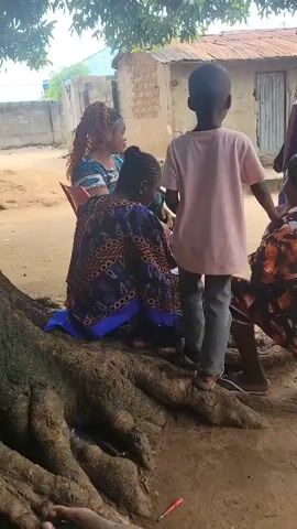 The video shows a group of five people gathered outdoors. In the foreground, a young child wearing a pink t-shirt and gray pants stands facing away from the camera, with their hands on the shoulders of a person seated on the ground. To the left, a woman with braided hair and a patterned blue and gold dress sits on a chair. To the right, a woman wearing an orange and black floral patterned dress and a black headscarf sits on a small wooden stool, also facing the child. Behind the child, to the right, a man in a maroon t-shirt stands and appears to be talking to the seated women. Behind the woman in blue, a woman with red hair is visible. The background consists of a rustic, possibly rural, setting with simple buildings and trees. The ground is dirt.