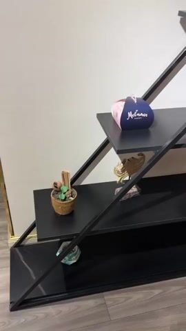 A black metal shelf unit with black shelves is against a white wall. On the shelves are various decorative items including a small plant in a pot, a pink and blue bag, a small drum, a model of the Eiffel Tower, and a ship in a bottle. To the right of the shelf unit, a mirror reflects a room with wooden floors, a metal railing, and a red chair.