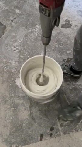 A person wearing gloves and grey sneakers is mixing a white liquid in a white bucket with a power mixer. The mixer is grey and red. The bucket is on a concrete floor. There is some white liquid splashed on the floor around the bucket.