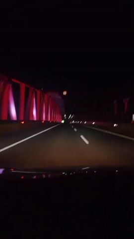The video shows a first-person perspective from inside a vehicle traveling on a highway at night. The highway is illuminated by red and pink lights lining the left side of the road, possibly from a bridge or overpass structure. The road itself has white lane markings and the headlights of the vehicle are on, casting a beam ahead. In the distance, other vehicle lights can be seen. The scene appears to be a highway tunnel or a section of highway with overhead illumination.
