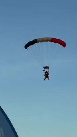 A tandem skydiver descends towards a grassy landing area with trees in the background. The skydiver, wearing a blue jumpsuit and a black helmet, is attached to an instructor who is also wearing a black helmet and a dark colored jumpsuit. The parachute is red, black, and tan and has lines connecting it to the skydivers. A large blue banner with a white logo is partially visible in the foreground. The sky is clear and blue with a hint of sunset colors. The skydivers are angled as if nearing the end of their descent.