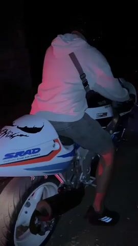 The video shows a person sitting on a Suzuki motorcycle at night. The person is wearing a white hoodie, gray shorts, black Adidas shoes, and a black helmet. The motorcycle is white with blue and orange stripes and has the words "SRAD Suzuki Ram Air Direct" on the side. The person revs the engine of the motorcycle, and the taillight illuminates. The setting appears to be outdoors in a dark environment.