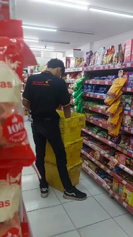 A young man wearing a black t-shirt with a logo and black pants is standing in a convenience store aisle. He is reaching into a stack of three yellow plastic crates. Shelves filled with snacks and other products are visible on the right side of the frame. The store appears to be well-lit with fluorescent lighting.