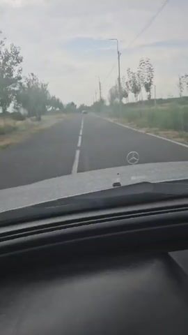 This is a first-person view from inside a vehicle traveling on a paved road. The road has a dashed white line down the center. Trees line both sides of the road, and there are power lines and streetlights visible in the distance. The sky is cloudy. The hood of the vehicle, featuring a Mercedes-Benz emblem, is visible in the foreground. Another vehicle is visible in the distance.