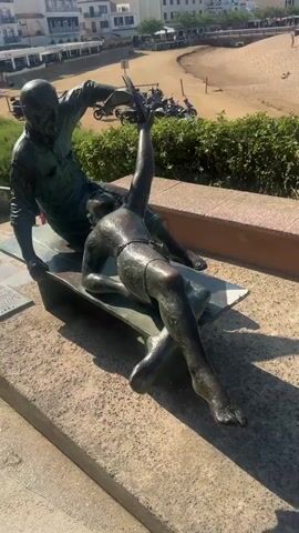 The video shows a bronze sculpture of two people sitting on a bench. One person is an older man with a bald head, wearing a shirt and pants, and holding a book. The other person is a younger person, possibly a woman, lying down with their head on the older man's lap. They are wearing shorts and have their arm raised. The sculpture is positioned on a stone ledge overlooking a beach with many people, umbrellas, and buildings in the background. The sea and a few boats are also visible. The weather appears to be sunny.