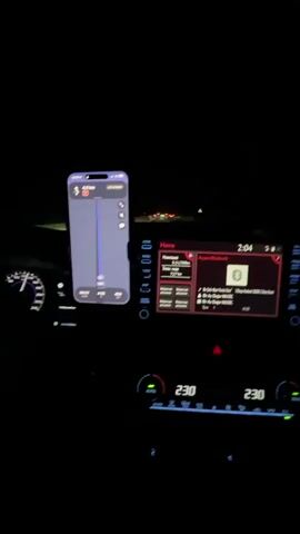 The video shows the interior of a car at night. The driver's side dashboard is visible, with a speedometer on the left. In the center, a smartphone is mounted and displays a navigation app, showing a route with distance and time information. To the right of the phone, the car's infotainment system screen is visible, displaying "Home" and "Bluetooth" information, including song title and artist. The time on the infotainment screen reads 2:04. Below the infotainment screen are climate control displays showing 23.0 degrees Celsius on both sides. The scene is dark, indicating it is nighttime, and the car appears to be in motion, with other vehicles visible in the distance on the road ahead.