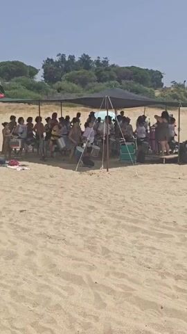A group of children and adults are gathered on a sandy beach under a large canopy. They are mostly sitting on chairs or the sand, facing a line of drums. A flag with the word "TUCURUCUM" is visible to the left of the canopy. The participants appear to be involved in a drumming activity. The environment is sunny with some trees in the background.