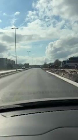 The view from inside a car driving down a paved road on a partly cloudy day. There are streetlights lining the road, and buildings visible on the left. Other cars are visible on the road and parked on the side. The sky is blue with large, puffy white clouds. The road appears to be in good condition. Construction or landscaping is taking place on the right side of the road. The car's dashboard is visible at the bottom of the frame.