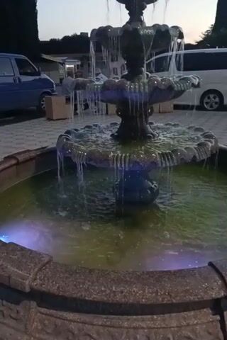 A video captures a close-up, low-angle shot of a multi-tiered fountain with water cascading down its levels. The fountain has an ornate, dark-colored structure with what appear to be decorative figures. The water in the basin of the fountain is illuminated by colored lights, transitioning from green to pink hues. In the background, a blue car is parked, and behind it, a white van is visible. The ground around the fountain is paved with light-colored interlocking bricks. Trees line the left side of the frame, and the sky, appearing to be late afternoon or early evening, is visible in the upper left corner. There are no visible people in the frame.