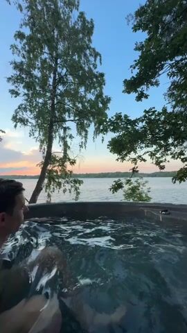 A man is in a hot tub by a lake at sunset. He is looking out at the water and trees. The water is bubbly and appears to be warm. He is wearing a dark bathing suit. The sun is setting in the distance, casting a warm glow on the sky.