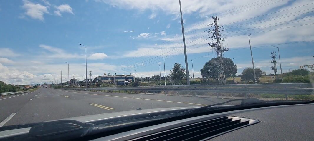 A car is driving down a highway under a blue sky with scattered white clouds. There is a McDonald's visible on the other side of the barrier. The road is fairly empty, with few other vehicles in sight. Power lines stretch across the sky.  A voice that sounds like a child can be heard.