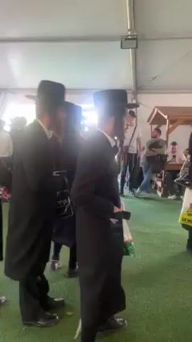 The video shows a group of Orthodox Jewish men in black hats and coats standing inside a tent. They are on artificial turf. Some of them are holding shopping bags. Other people are seated at tables in the background. The lighting is bright.