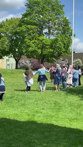 A group of people of various ages and ethnicities are dancing in a circle on a grassy field. They are holding hands and moving in a circular motion. Some are wearing headscarves. There are trees, a building, and a flagpole in the background. The sky is blue with some clouds. The scene appears to be outdoors on a sunny day. There is a person sitting on a bench in the bottom left corner of the frame, partially obscured.