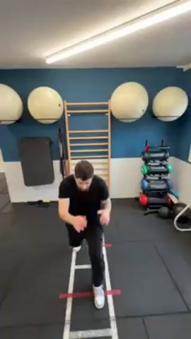 The video shows a man working out. He is using an agility ladder on the floor, stepping in and out of the rungs quickly. He is also throwing punches as he steps. He is wearing a black shirt and black pants. The workout space has black floors and a blue and white wall. There is a ladder, exercise balls, weights, and other exercise equipment in the background.
