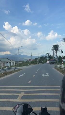 The video shows a view from a moving motorcycle driving on a paved road in a rural area. The road has yellow dashed lines dividing the lanes. On either side of the road are patches of green vegetation and some buildings. In the background, there are mountains visible beneath a partly cloudy sky. The clouds are a mix of white and gray, indicating potential for rain. Power lines and utility poles run alongside the road. A truck is parked on the side of the road ahead, and another vehicle can be seen further away. The camera is pointed forward, capturing the motorcycle's perspective.